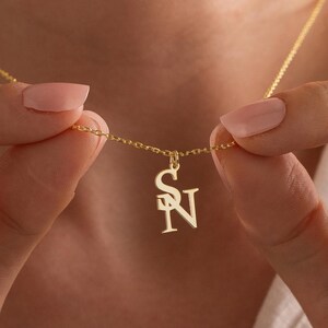 Puede incluir: Un collar dorado con un colgante con las letras entrelazadas "S" y "N". La cadena está sujeta por una persona. El colgante mide aproximadamente 2 cm de altura. El collar es una joya.