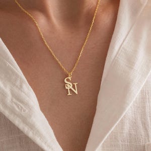 Puede incluir: Un collar dorado con una cadena delicada y un colgante con las letras entrelazadas "S" y "N". El collar se muestra sobre una camisa blanca abotonada.