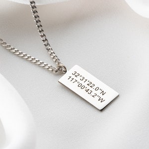 May include: A silver necklace featuring a rectangular pendant with engraved coordinates: "32°31'22.0"N 117°00'43.2"W". The chain is a curb style. The background is a white fabric with a pink flower.