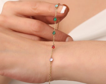 May include: A gold chain bracelet with five colorful gemstones. The gemstones are green, blue, red, and purple.
