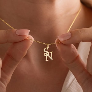 May include: A gold-colored necklace with a pendant featuring the intertwined letters "S" and "N". The delicate chain is held by a person. The pendant is approximately 0.75 inches in height. The necklace is a piece of jewelry.
