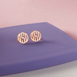 May include: A pair of gold-toned monogram earrings with a circular design. The earrings are on a purple surface.