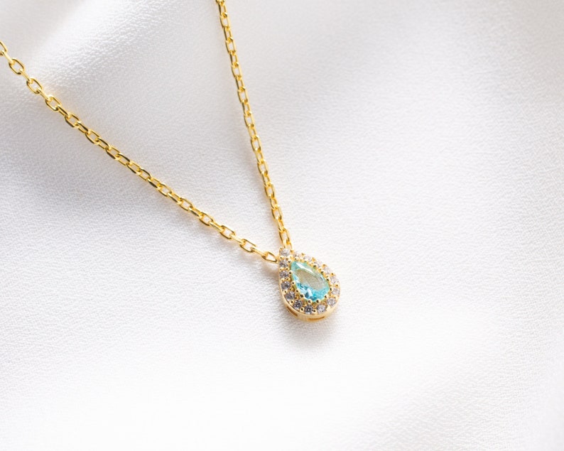 May include: A delicate gold chain necklace with a teardrop-shaped pendant featuring a light blue gemstone surrounded by a halo of smaller clear stones.