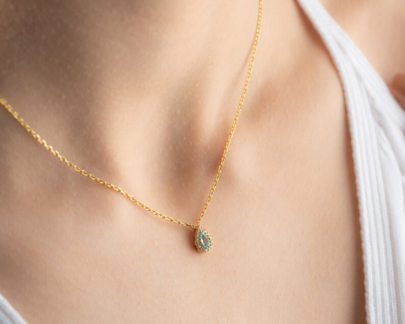 May include: A gold chain necklace with a teardrop-shaped pendant featuring a light blue gemstone surrounded by smaller clear gemstones.