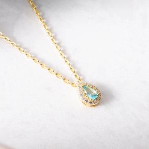 May include: A gold chain necklace with a teardrop-shaped pendant featuring a light blue gemstone surrounded by smaller clear gemstones.