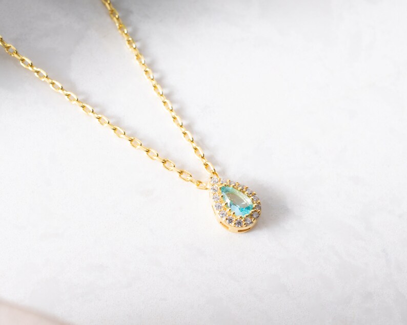 May include: A gold chain necklace with a teardrop-shaped pendant featuring a light blue gemstone surrounded by smaller clear gemstones.