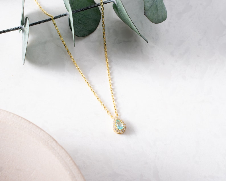 May include: A delicate gold chain necklace with a teardrop-shaped pendant featuring a light blue gemstone surrounded by smaller clear stones.