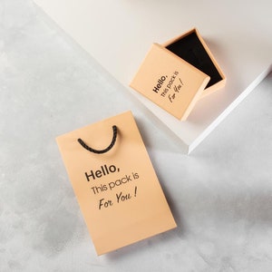 May include: A light brown gift bag with black handles and the text "Hello, This pack is For You!" printed on it. A smaller light brown box with the same text is also shown.