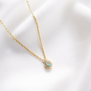 May include: A delicate gold chain necklace with a teardrop-shaped pendant. The pendant is set with a light blue gemstone surrounded by smaller clear gemstones.