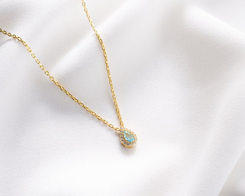 May include: A delicate gold chain necklace with a teardrop-shaped pendant. The pendant is set with a light blue gemstone surrounded by smaller clear gemstones.