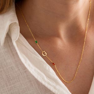 May include: A gold-colored necklace with a delicate chain. The necklace features a capital letter "G" pendant, along with a green and a red rectangular gemstone. The necklace is worn against a white shirt.