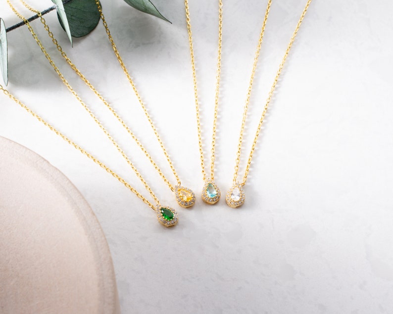 May include: Four gold necklaces with teardrop-shaped pendants. Each pendant is set with a different coloured gemstone: green, blue, light blue, and clear.