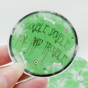 May include: A green and white round sticker with the text "Double Double, Toil and Trouble" in a glittery font. The sticker has a white border and a white background with a green circle in the centre.