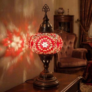 Puede incluir: Una lámpara de mesa con una pantalla esférica de vidrio compuesta por azulejos de mosaico rojos y blancos dispuestos en forma de estrella. La lámpara tiene una base de metal color bronce oscuro y un remate decorativo que proyecta luz roja.