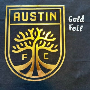 May include: Black fabric with a gold foil emblem. The emblem features the word "AUSTIN" at the top, with a tree design and the letters "F" and "C" at the bottom. The words "Gold Foil" are written in white.