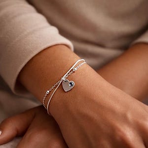 May include: A silver bracelet featuring a heart-shaped charm and a small blue gemstone. The bracelet is worn on a wrist, with a second, delicate chain with small beads. The background is a neutral colour.