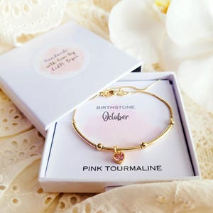 K&ouml;nnte beinhalten: Ein goldfarbenes Armband mit einem rosa herzf&ouml;rmigen Anh&auml;nger, pr&auml;sentiert in einer wei&szlig;en Geschenkbox. Die Box enth&auml;lt eine Karte mit der Aufschrift "Geburtsstein Oktober Pinker Turmalin". Der Deckel der Box tr&auml;gt die Aufschrift "Handmade with love by Kidi Bips".