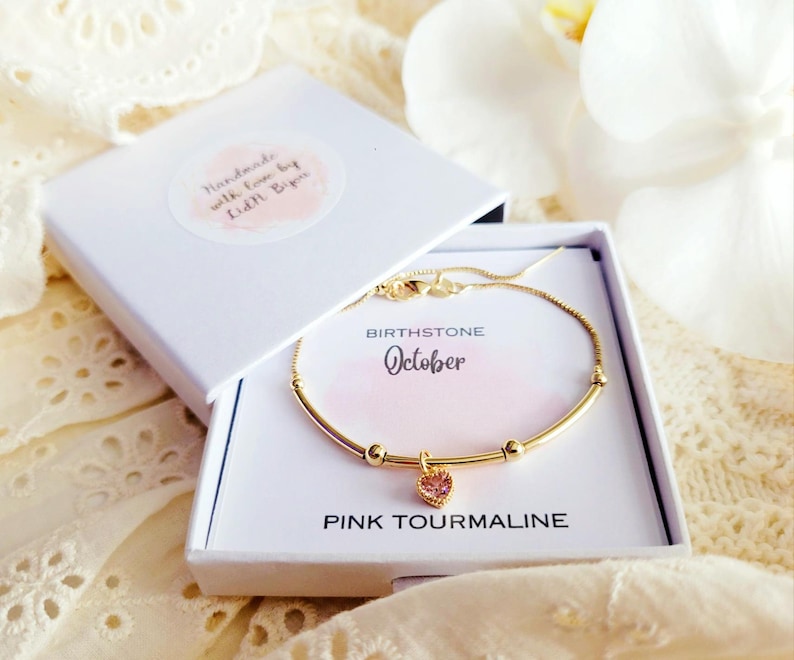 K&ouml;nnte beinhalten: Ein goldfarbenes Armband mit einem rosa herzf&ouml;rmigen Anh&auml;nger, pr&auml;sentiert in einer wei&szlig;en Geschenkbox. Die Box enth&auml;lt eine Karte mit der Aufschrift "Geburtsstein Oktober Pinker Turmalin". Der Deckel der Box tr&auml;gt die Aufschrift "Handmade with love by Kidi Bips".