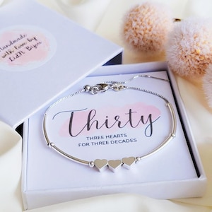 May include: A silver bracelet with three heart charms inside a white gift box. The box contains a card with the word "Thirty" and the phrase "Three Hearts for Three Decades". The box is open, revealing the bracelet.