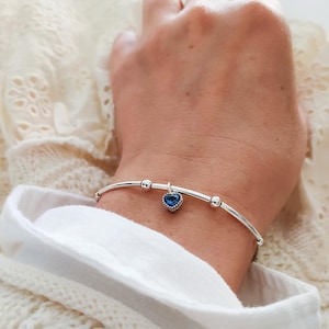 K&ouml;nnte beinhalten: Ein silberfarbenes Armband mit einem herzf&ouml;rmigen blauen Anh&auml;nger. Das Armband hat ein geschwungenes Design mit kleinen, runden Verzierungen. Der Anh&auml;nger ist tiefblau und mit kleinen, klaren Steinen besetzt. Das Armband wird am Handgelenk getragen.