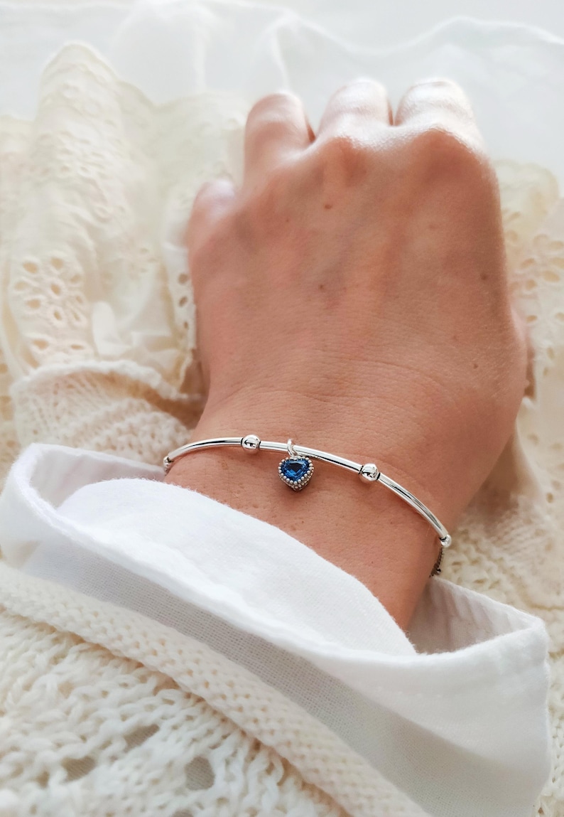 K&ouml;nnte beinhalten: Ein silberfarbenes Armband mit einem herzf&ouml;rmigen blauen Anh&auml;nger. Das Armband hat ein geschwungenes Design mit kleinen, runden Verzierungen. Der Anh&auml;nger ist tiefblau und mit kleinen, klaren Steinen besetzt. Das Armband wird am Handgelenk getragen.
