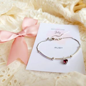 K&ouml;nnte beinhalten: Ein silbernes Armband mit einem herzf&ouml;rmigen Rubin-Anh&auml;nger, pr&auml;sentiert auf einer Karte mit der Aufschrift "Ruby" und "July Birthstone". Eine rosa Satinschleife ist links gebunden. Das Armband liegt auf einer cremefarbenen, strukturierten Oberfl&auml;che.