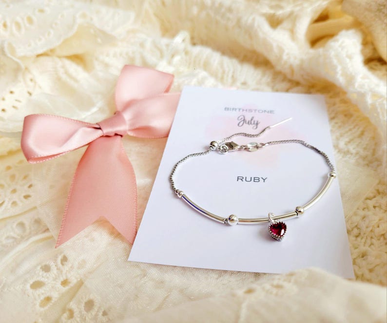 K&ouml;nnte beinhalten: Ein silbernes Armband mit einem herzf&ouml;rmigen Rubin-Anh&auml;nger, pr&auml;sentiert auf einer Karte mit der Aufschrift "Ruby" und "July Birthstone". Eine rosa Satinschleife ist links gebunden. Das Armband liegt auf einer cremefarbenen, strukturierten Oberfl&auml;che.
