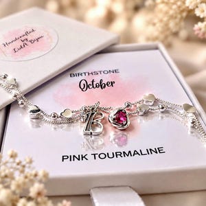 May include: A silver-toned bracelet featuring small heart-shaped charms, a letter B charm, and a pink tourmaline stone. The bracelet rests on a white card labelled BIRTHSTONE October and PINK TOURMALINE, inside a white box with a LidA Bijou sticker.