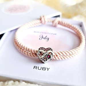 May include: A pale pink rope bracelet with a heart-shaped charm, displayed in a white gift box. The charm is silver-toned with red details and an infinity symbol. The box includes the words "July" and "Ruby".