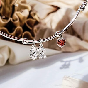 May include: A silver bracelet with dangling charms. The charms include the number "32" in a sparkling, clear stone design, and a small heart-shaped charm with a red stone. The bracelet has small silver beads.