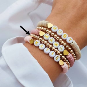 Könnte beinhalten: Ein Stapel Armbänder mit goldenen Perlen und weißen Buchstabenperlen, die die Namen "Katie", "Mama" und "Ruby" buchstabieren. Die Armbänder befinden sich am Handgelenk einer Person.