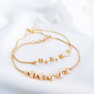 May include: Two gold-coloured bracelets. One bracelet has the word "MAMA" spelled out in gold letters on clear beads. The other bracelet has gold heart-shaped charms. Both bracelets are on a white background.