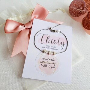 May include: A silver chain bracelet with three star charms. The bracelet is displayed on a white card with the text "Thirty Three Stars for Three Decades" and "Handmade with love by Lid'H Bijou".