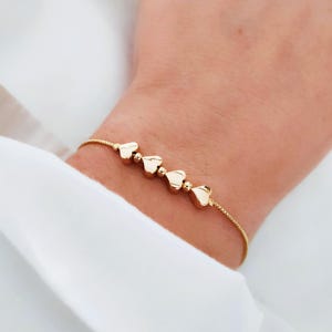 May include: A gold bracelet with heart-shaped charms. The bracelet is worn on a wrist, with a white shirt in the background. The bracelet's chain is thin, and the hearts are evenly spaced.