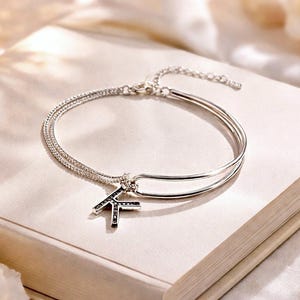 Peut inclure: Un bracelet argenté à double rangée avec un charm en forme de lettre "K". Le charm est orné de petites pierres sombres. Le bracelet est présenté sur une surface de couleur claire.