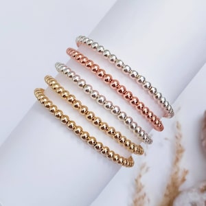 Könnte beinhalten: Vier Perlenarmbänder in Gold, Silber und Roségold. Die Armbänder sind übereinander gestapelt.