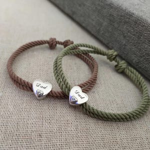 May include: Two rope bracelets, one in brown and one in green, each with a silver heart charm. The heart charms are engraved with the word "Dad". The bracelets are knotted and lie on a neutral fabric.