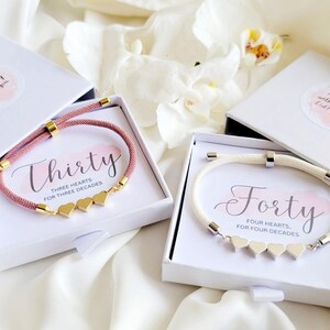 May include: Two bracelets in white boxes. One is a pink cord bracelet with gold accents and three heart charms. The other is a white cord bracelet with silver accents and four heart charms. The boxes have the words "Thirty" and "Forty" on the cards.