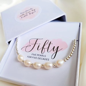May include: A white box containing a silver bracelet with five white pearls and silver beads. The box has a pink and white label that reads "Fifty" and "Five Pearls for Five Decades".