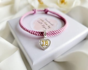 May include: A pink braided cord bracelet with a silver heart charm that has the number 18 in gold lettering and clear rhinestones around the edge.