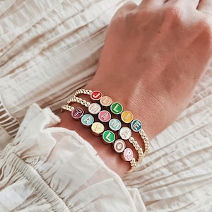 May include: Gold beaded bracelets with colourful round beads spelling out names. The names "JULIE", "MAMA", "TAYLOR", and "LOR" are visible. The bracelets are worn on a wrist against a cream-coloured background.