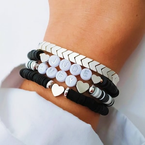 May include: A stack of bracelets with black and white beads, silver arrow charms, and white letter beads spelling "JUDE RUBY".