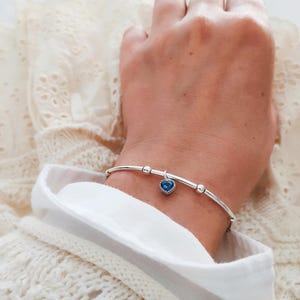 K&ouml;nnte beinhalten: Ein silbernes Armband mit einem blauen herzf&ouml;rmigen Anh&auml;nger. Das Armband hat ein schlankes Design mit kleinen silbernen Perlen. Der Herzanh&auml;nger ist leuchtend blau und setzt einen Farbakzent. Das Armband wird am Handgelenk getragen.