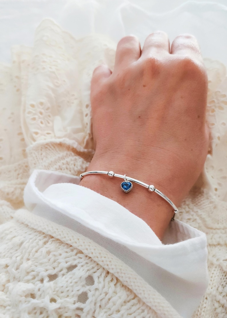 K&ouml;nnte beinhalten: Ein silbernes Armband mit einem blauen herzf&ouml;rmigen Anh&auml;nger. Das Armband hat ein schlankes Design mit kleinen silbernen Perlen. Der Herzanh&auml;nger ist leuchtend blau und setzt einen Farbakzent. Das Armband wird am Handgelenk getragen.