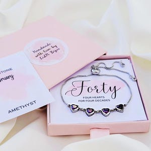 Könnte beinhalten: Ein silbernes Armband mit vier herzförmigen Amethyststeinen, präsentiert in einer rosa Geschenkbox. Die Box enthält eine Karte mit der Aufschrift "BIRTHSTONE February AMETHYST" und einen Aufkleber "Handmade with love by Lidl Biyou". Auf dem Armband steht das Wort "Forty".