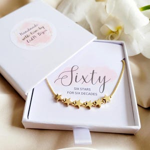 May include: A white box with a gold necklace inside. The necklace has six gold stars and the text "Sixty Six Stars for Six Decades". The box has a pink and white label with the text "Sixty" in cursive.
