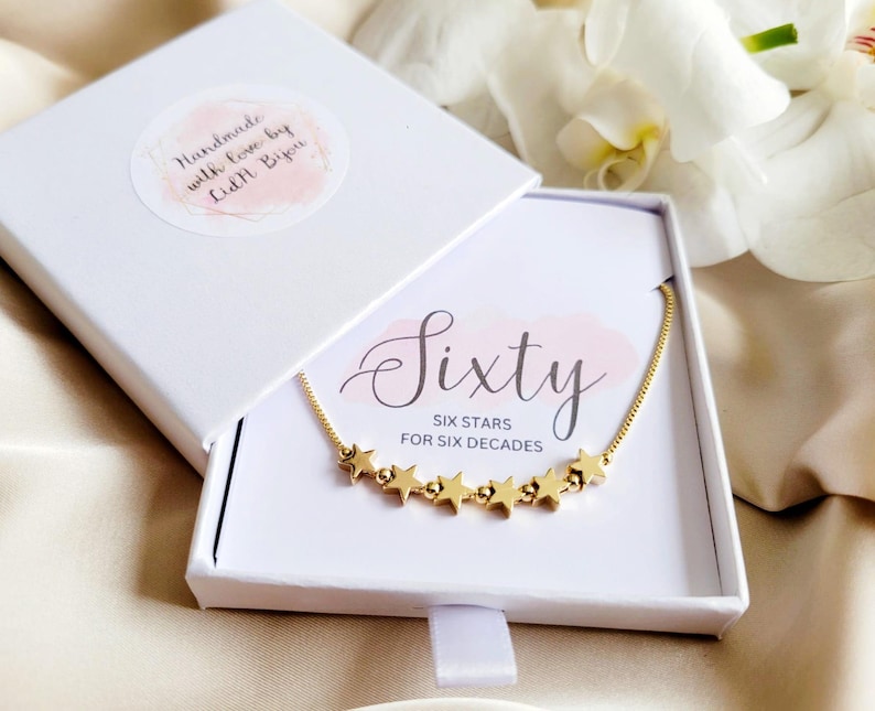 May include: A white box with a gold necklace inside. The necklace has six gold stars and the text "Sixty Six Stars for Six Decades". The box has a pink and white label with the text "Sixty" in cursive.