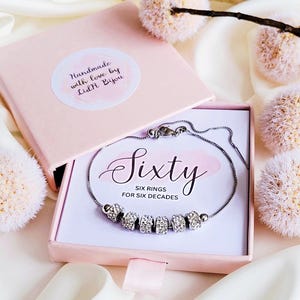 May include: A silver bracelet with six rhinestone-studded rings is displayed in a pink gift box. The box is open, revealing the bracelet against a white background with the word "Sixty" and the phrase "Six Rings for Six Decades". The box is surrounded by decorative flowers.