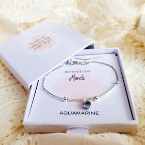 K&ouml;nnte beinhalten: Ein silbernes Armband mit einem herzf&ouml;rmigen Aquamarin-Edelstein, pr&auml;sentiert in einer offenen wei&szlig;en Geschenkbox. Die Box tr&auml;gt die Aufschrift "BIRTHSTONE March AQUAMARINE". Die Box hat einen kleinen Aufkleber mit der Aufschrift "Handmade with love by Edith Bipin".