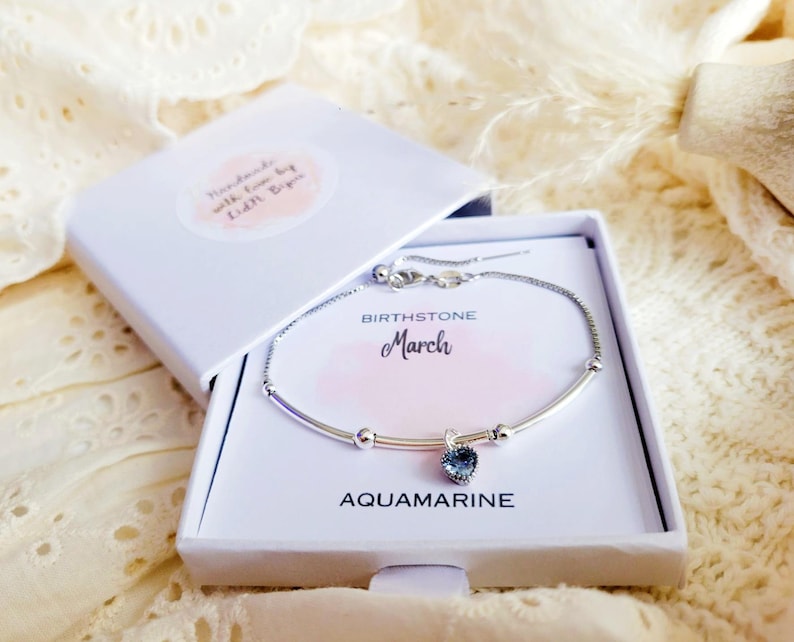 K&ouml;nnte beinhalten: Ein silbernes Armband mit einem herzf&ouml;rmigen Aquamarin-Edelstein, pr&auml;sentiert in einer offenen wei&szlig;en Geschenkbox. Die Box tr&auml;gt die Aufschrift "BIRTHSTONE March AQUAMARINE". Die Box hat einen kleinen Aufkleber mit der Aufschrift "Handmade with love by Edith Bipin".